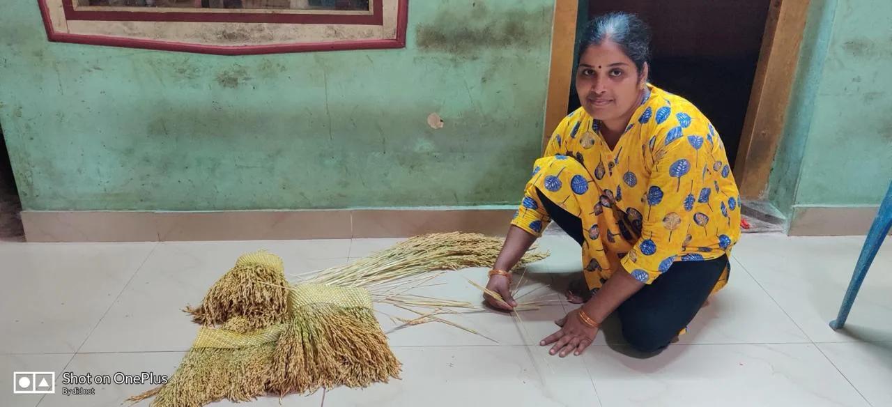 A woman artisan kneeling beside bundles of dried paddy and a half-made toran